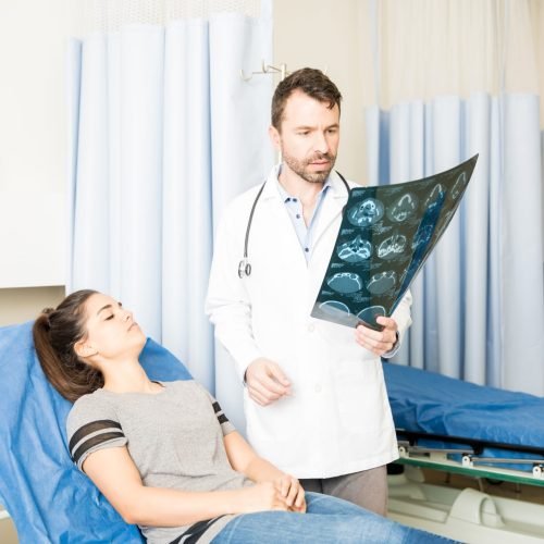 Handsome hispanic doctor looking at CT scan results of a female patient lying on bed in emergency room